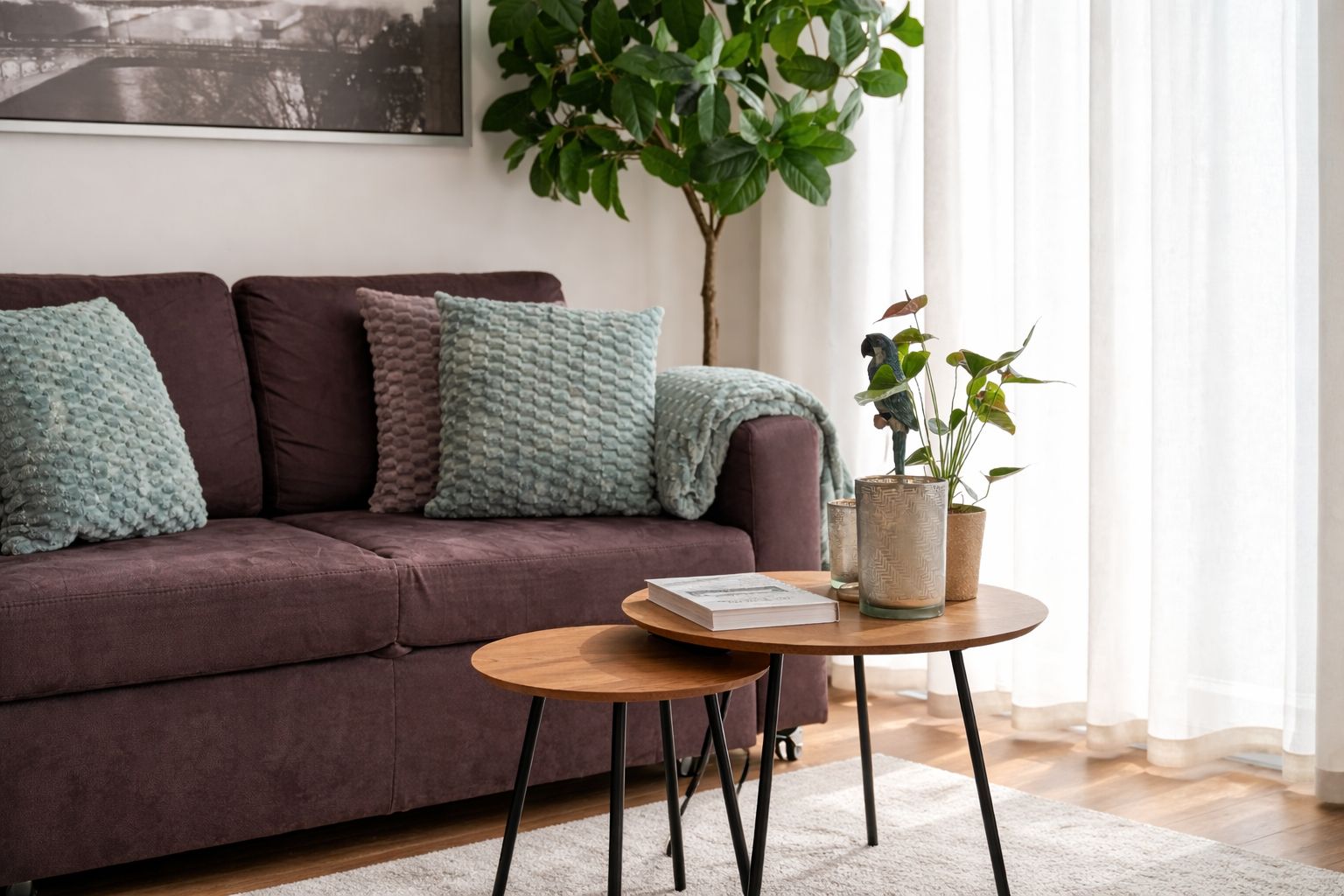 Ein Wohnzimmer mit einer Couch, Kissen, einem runden Tisch mit einem Buch, einer Pflanze und einem Fenster mit Vorhängen.