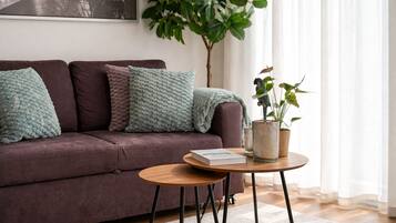 Ein Wohnzimmer mit einer Couch, Kissen, einem runden Tisch mit einem Buch, einer Pflanze und einem Fenster mit Vorhängen.