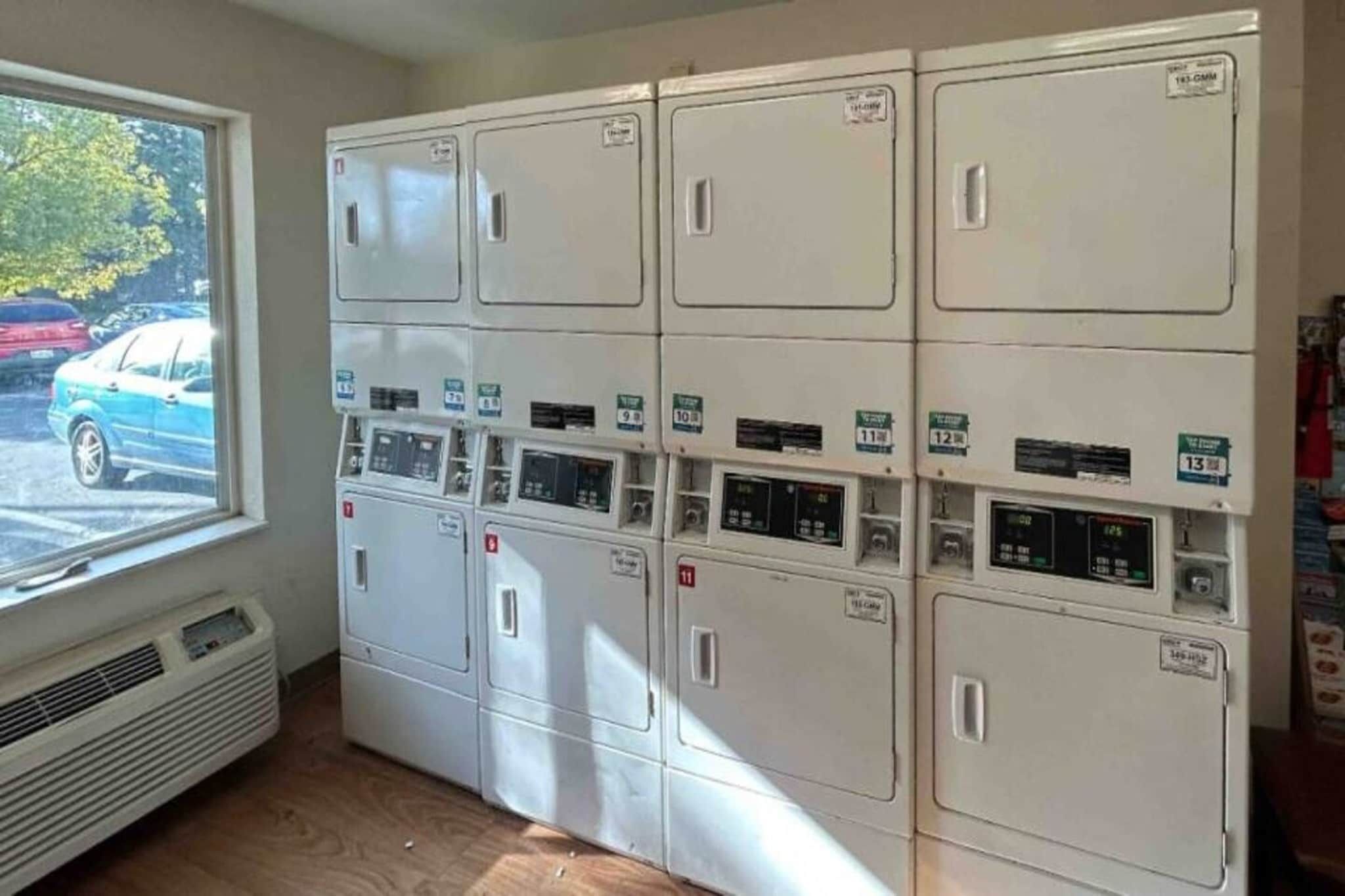 Une buanderie équipée de plusieurs machines à laver et de plusieurs séchoirs, une fenêtre donnant sur les voitures garées et une bouche d’aération dans le mur.