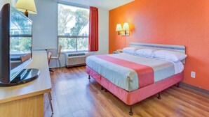 Habitación de hotel con cama, escritorio con lámpara, silla, ventana y unidad de aire acondicionado.