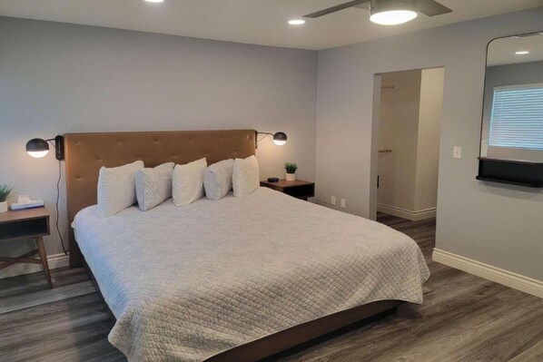 A bedroom with a large bed, a nightstand, a wall-mounted lamp, and a door leading to another room. - 1 Queen Bed | Premium, Mini Fridge (Las Vegas)