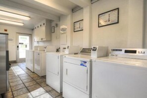 Une buanderie équipée de plusieurs machines à laver et de plusieurs séchoirs, avec un sol carrelé et une porte donnant sur une autre pièce.
