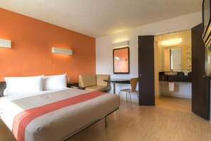 A hotel room with a bed, a desk, a chair, a mirror, and a bathroom. - 1 Full Bed | ADA/RI Shower (Las Vegas)