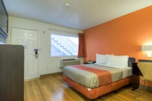 A hotel room with a bed, a desk, a chair, a window with a view of stairs, and a door. - 1 Full Bed | ADA/RI Shower (Las Vegas)