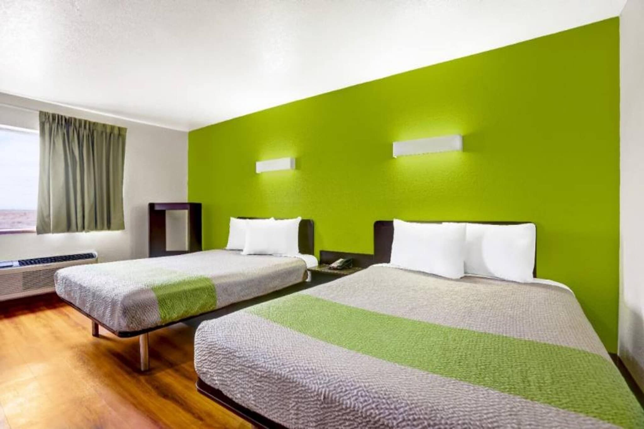 Hotellihuone, jossa on kaksi sänkyä, ikkuna verhoilla, lämmitin ja yksi seinä, joka on maalattu vihreällä korostusvärillä.