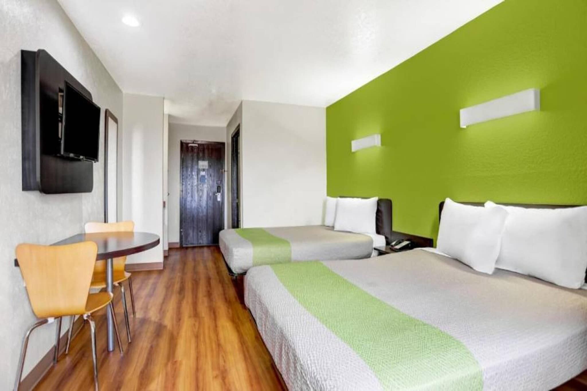 Habitación de hotel con piso de madera, una silla sencilla, una cama con ropa de cama blanca y verde, y un televisor fijado a la pared.