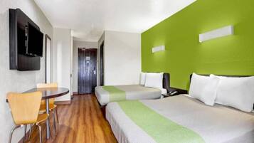 Habitación de hotel con piso de madera, una silla sencilla, una cama con ropa de cama blanca y verde, y un televisor fijado a la pared.