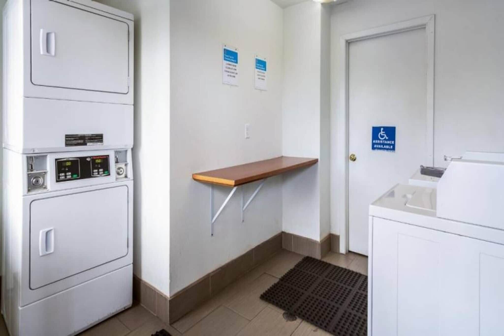 Quarto de hotel com máquina de lavar roupa, uma pequena estante de madeira, porta com identificação de acessibilidade e chão em azulejos.