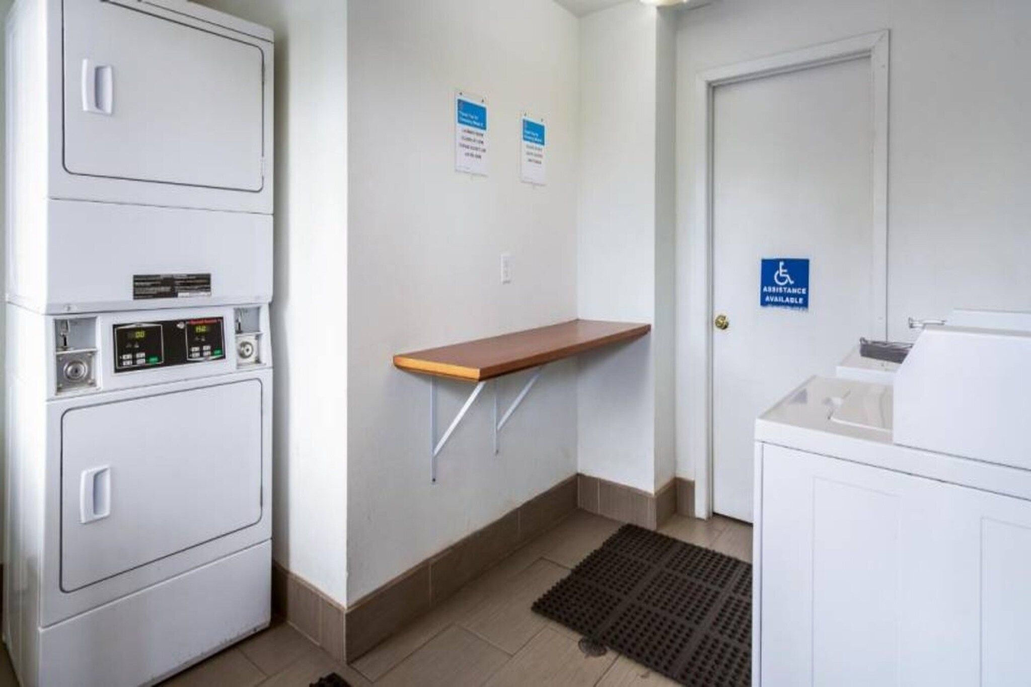 Quarto de hotel com um guarda-roupa branco, uma pequena prateleira de madeira, uma porta com o sinal de acessibilidade e piso de azulejos.