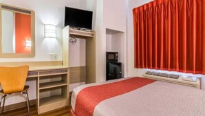 Una habitació d'hotel amb llit, escriptori, cadira, televisió, cafetera i finestra amb cortines.