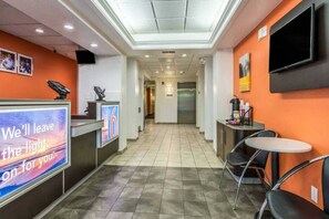 Um hall de hotel com uma mesa de recepção, uma televisão fixada na parede, uma área de estar com cadeiras e piso em azulejos.