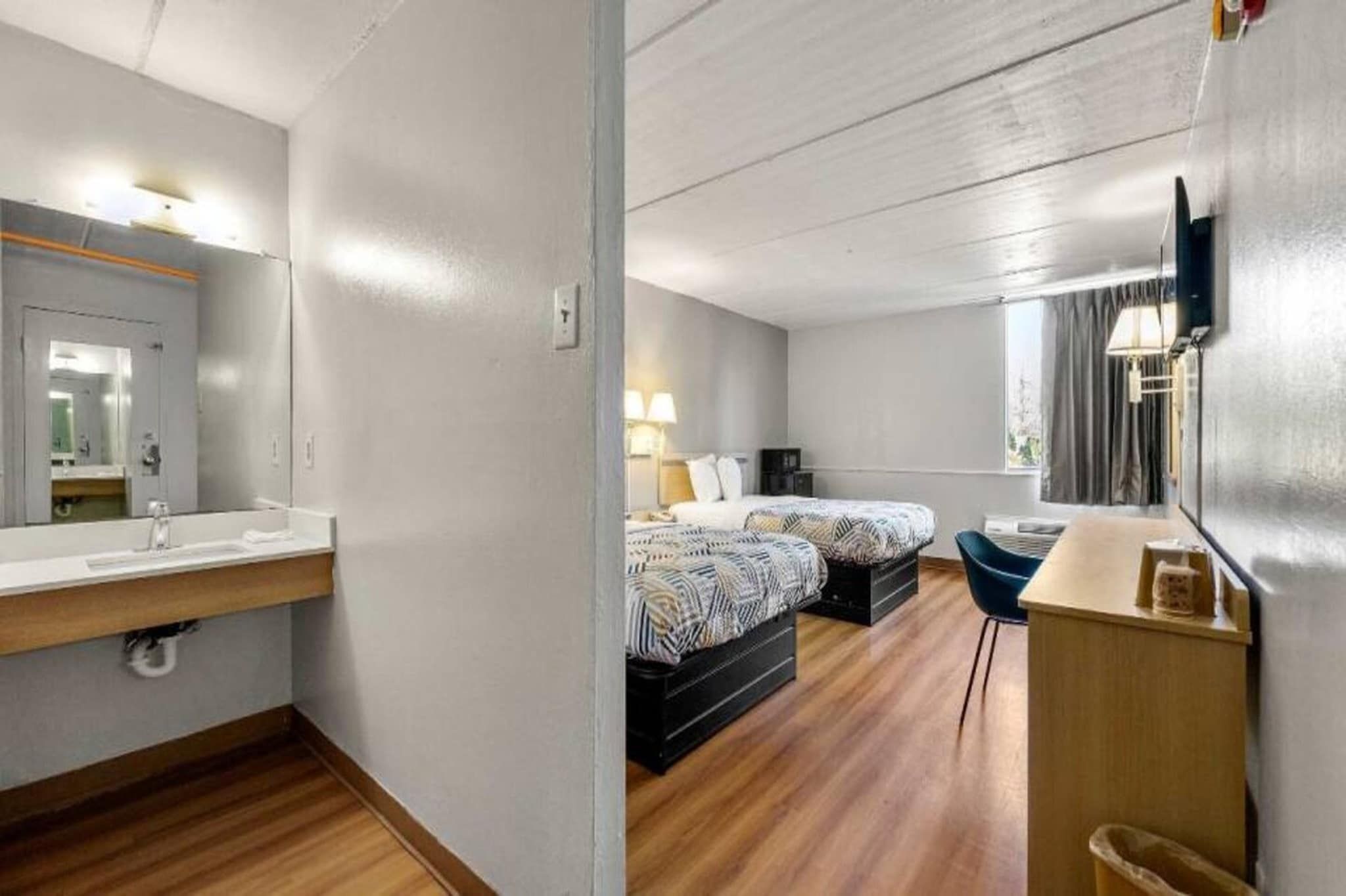 A hotel room with a wooden floor, a bed with patterned bedding, a desk with a chair, and a large mirror.