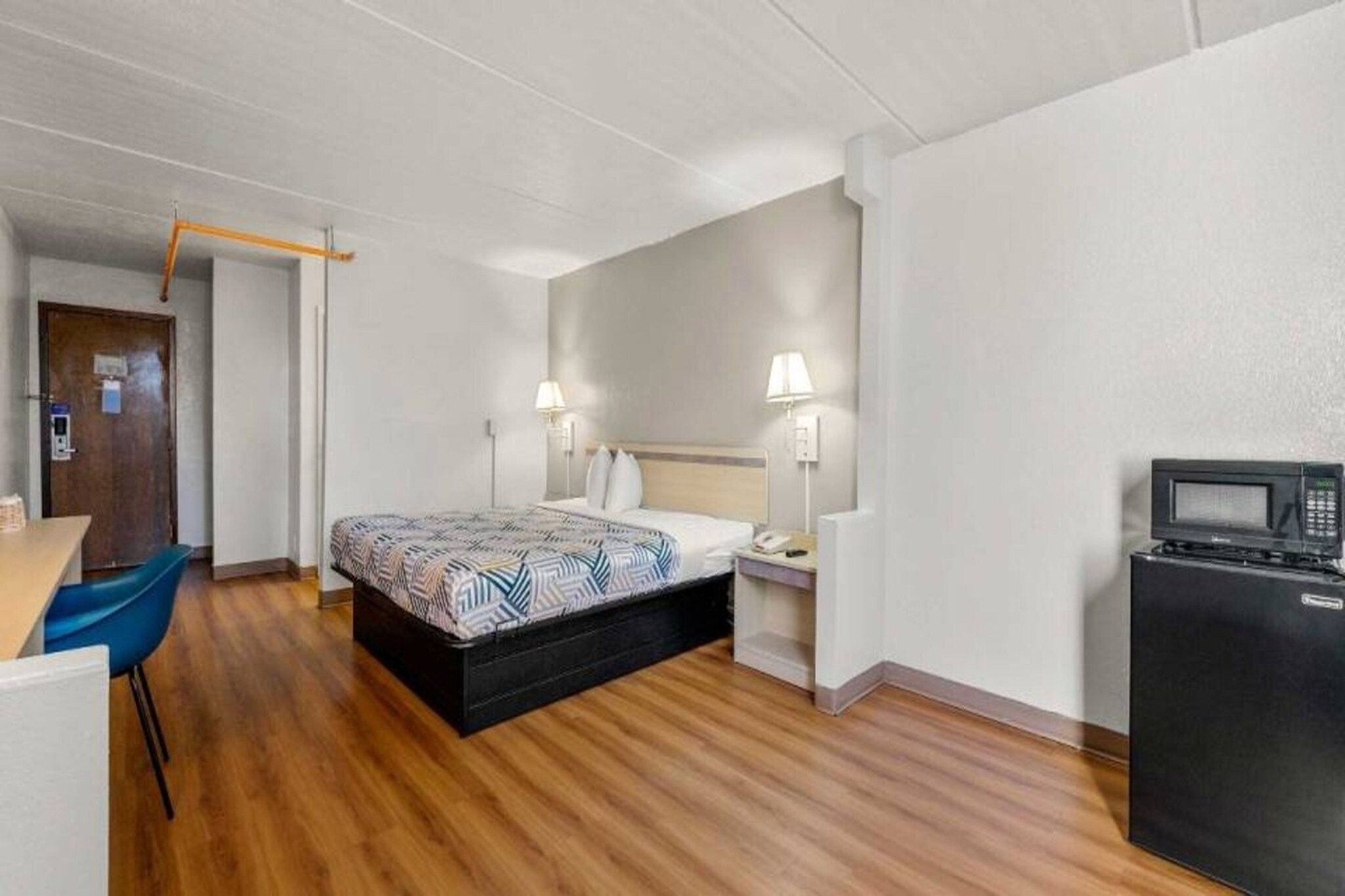 A hotel room with a bed, bedside table, lamp, desk, chair, and a refrigerator.