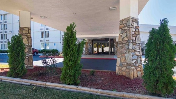 Entrada de hotel com galeria coberta, pilares de pedra e um estacionamento ao fundo.