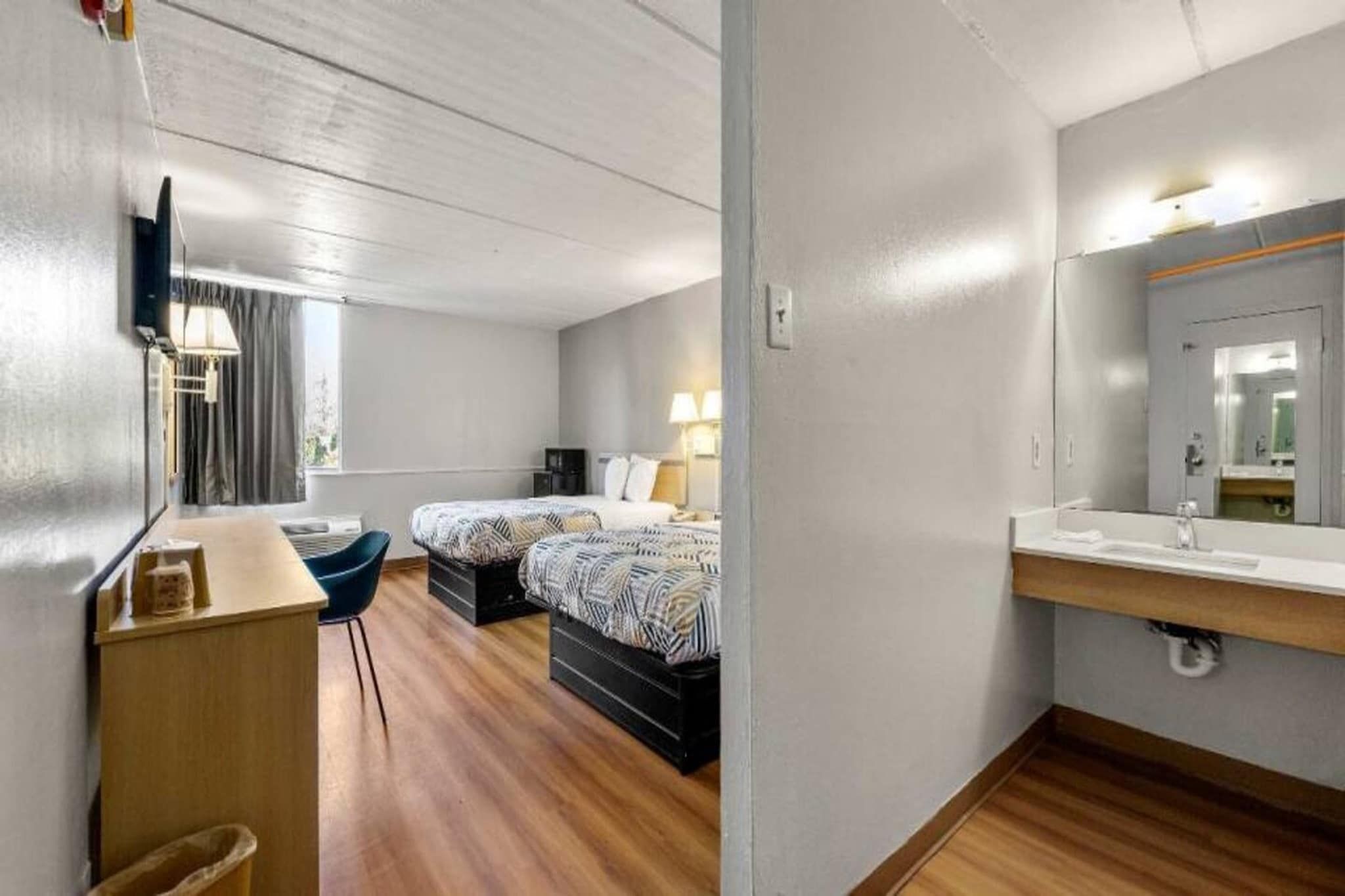 Habitación de hotel con cama, mesita de noche, lámpara y baño con espejo y lavamanos.