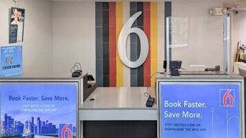 Een hotel receptiebalie met een groot nummer 6 op de muur, twee computerschermen en promotieposters voor boeken en besparen.