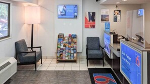 Un área de recepción con un mostrador, sillas, un atril para revistas y una pantalla digital.