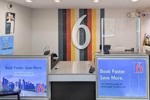 A hotel reception desk with a large '6' sign, a computer, and promotional posters for booking through the motel's website or app. - Spacious King Bed|Free Wifi|Vending (Nashua)