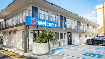 Een motel met een blauw bord waarop '6 Welcome' staat, een parkeerplaats met parkeerplaatsen voor mindervaliden, en een bloempotje op de voorgrond.