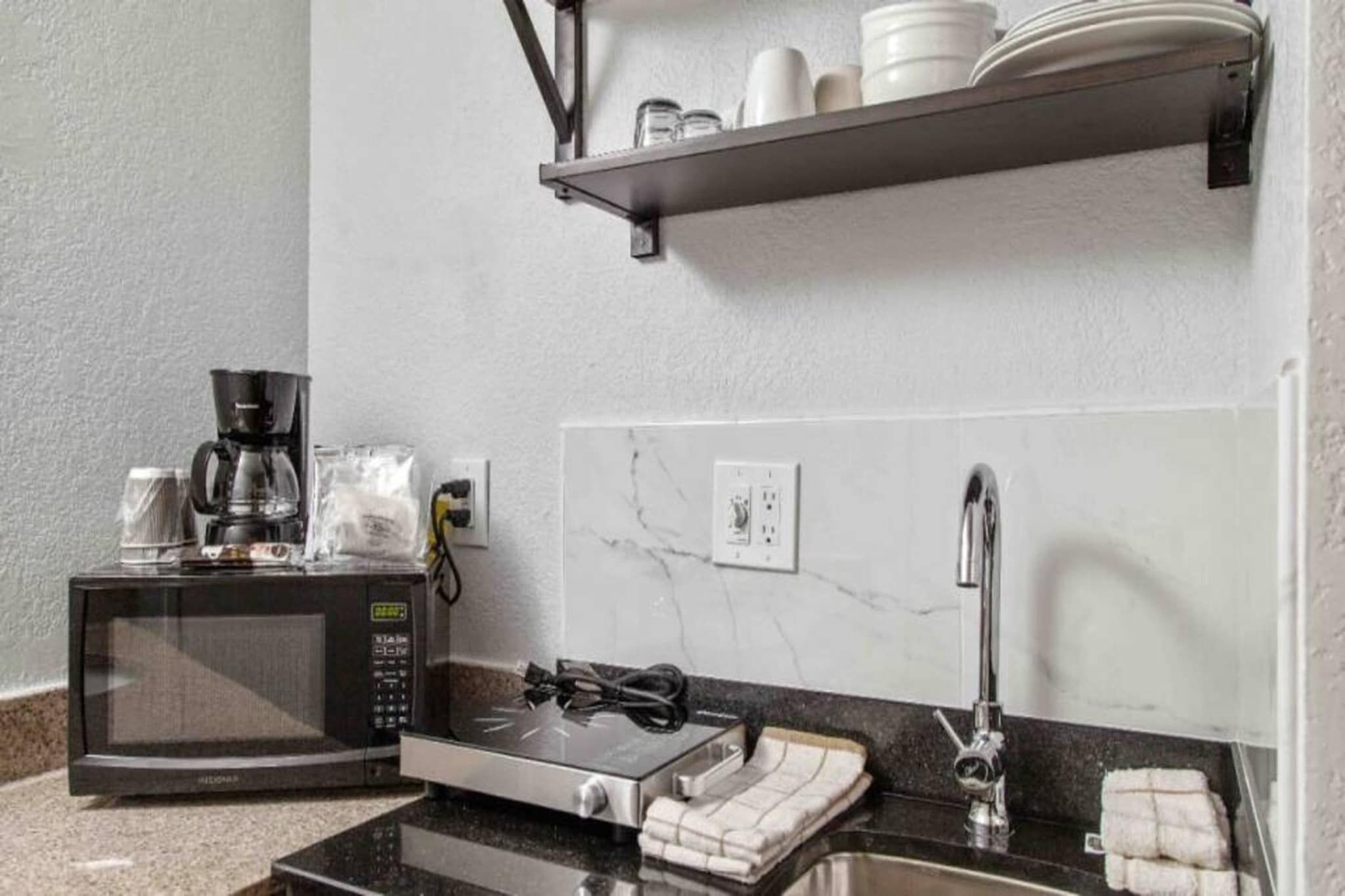 A compact kitchenette with a microwave, coffee maker, and a small oven. There is a sink with a faucet, and a shelf above holding plates and cups.