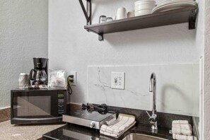 Une petite cuisine équipée d’un four à micro-ondes, d’une machine à café et d’un petit four. On y trouve un évier avec un robinet, et une étagère au-dessus pour ranger les assiettes et les tasses.