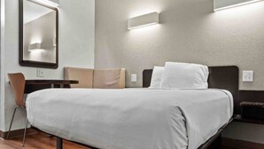Quarto de hotel com uma cama, uma cadeira, uma escrivaninha e um espelho.