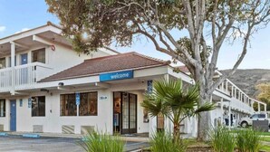 Um motel de dois andares com um letreiro de 'welcome', um estacionamento e uma árvore em primeiro plano.