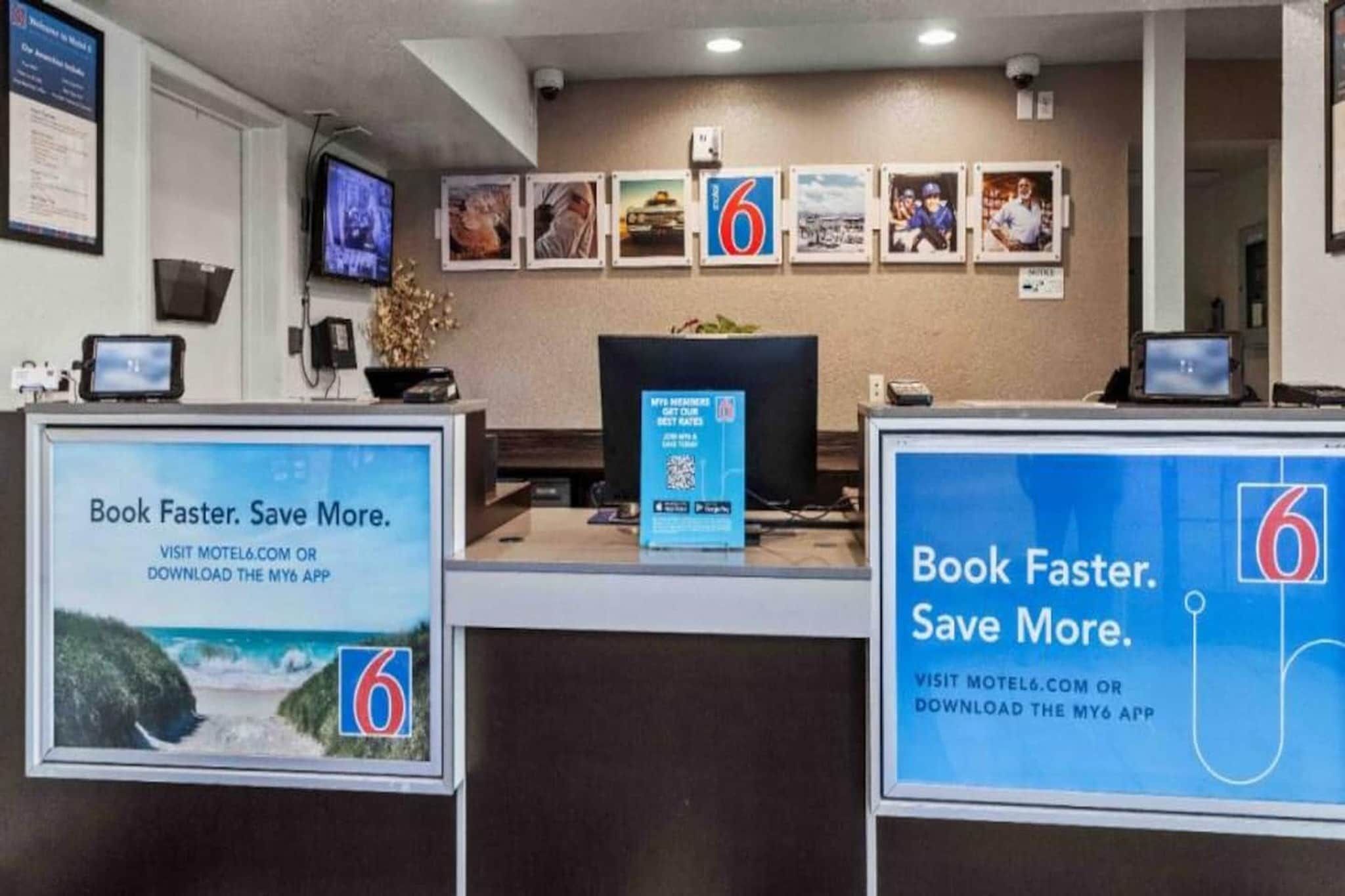 Bancada de recepção de hotel com monitor de computador, televisão e quadros na parede. Existem dois cartazes promocionais grandes em ambos os lados da bancada.