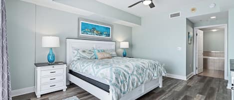 Un dormitorio con cama, mesitas de noche, ventilador de techo, un cuadro y una puerta que da a otra habitación.