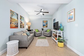 A living room with a grey sofa, a white side table, a floor lamp, a ceiling fan, and a TV stand with a flat-screen TV. - Spacious 3BR/3BA oceanfront condo in Daytona Beach Shores. Sleeps 8 with beautiful ocean views, a large private balcony, heated oceanfront pool and direct beach access. (Daytona Beach Shores)