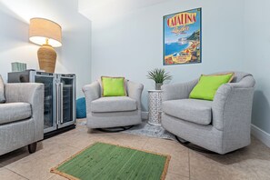 A living room with two grey armchairs, a green rug, a lamp, a refrigerator, and a poster on the wall. - Spacious 3BR/3BA oceanfront condo in Daytona Beach Shores. Sleeps 8 with beautiful ocean views, a large private balcony, heated oceanfront pool and direct beach access. (Daytona Beach Shores)