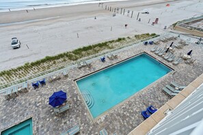 A pool area with lounge chairs and umbrellas, a beach in the background, and a white car parked on the sand. - Spacious 3BR/3BA oceanfront condo in Daytona Beach Shores. Sleeps 8 with beautiful ocean views, a large private balcony, heated oceanfront pool and direct beach access. (Daytona Beach Shores)