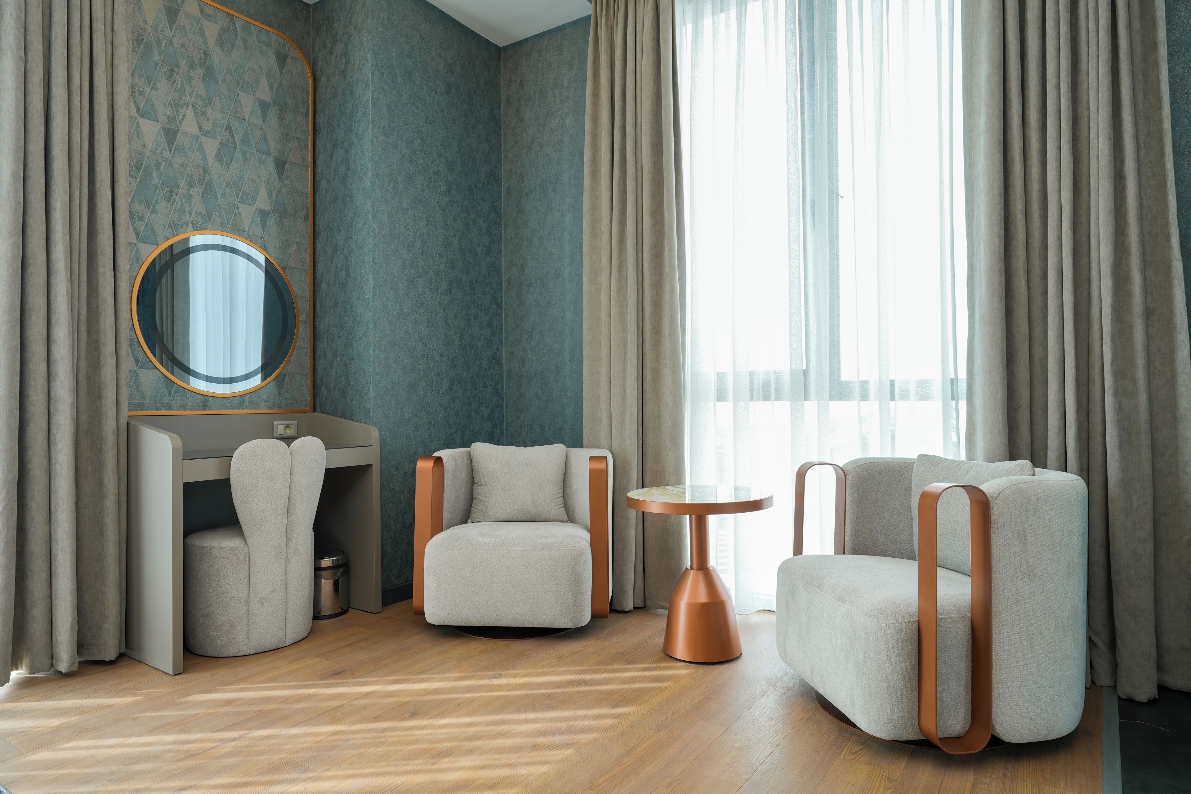 Ein modernes Zimmer mit Holzboden, einem runden Tisch, zwei Sesseln, einem Schreibtisch mit Spiegel und einem großen Fenster mit Vorhängen.