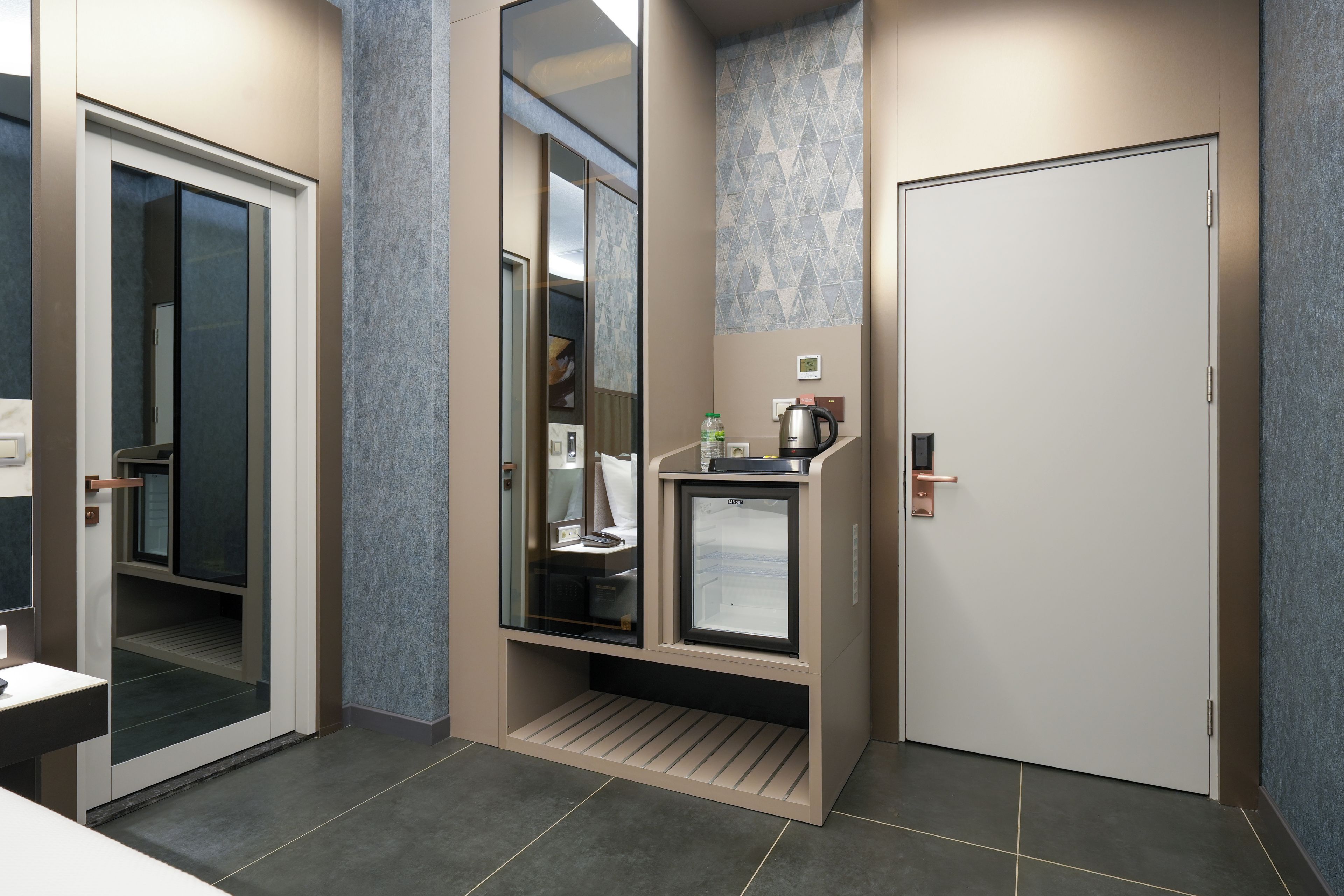 A modern hotel room with a minibar, a mirror, a door, and a small seating area.