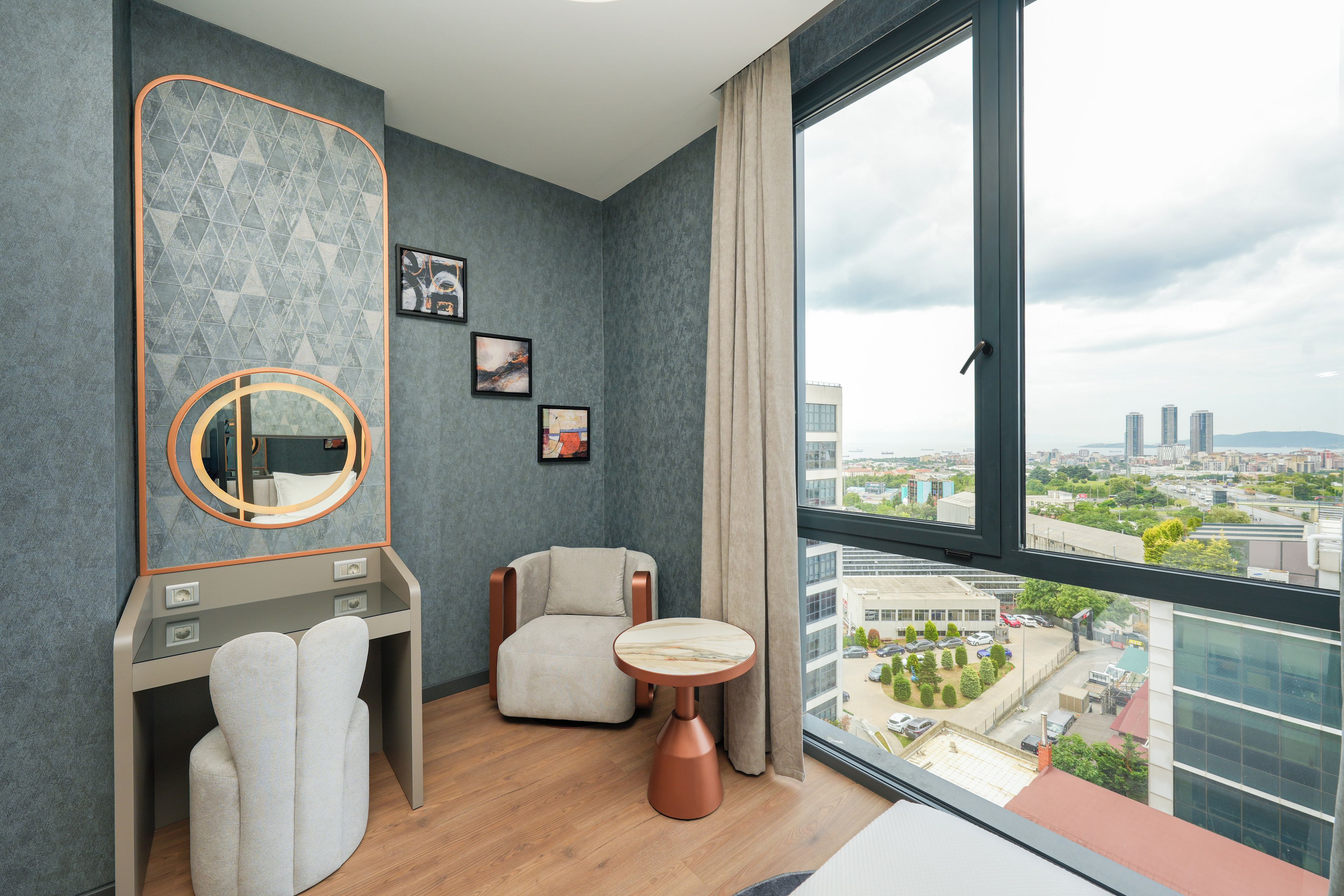 Una habitación de hotel moderna con un ventanal grande, un espejo redondo, una tocador con una toalla, una silla y una mesita.