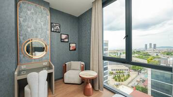 Una habitación de hotel moderna con un ventanal grande, un espejo redondo, una tocador con una toalla, una silla y una mesita.
