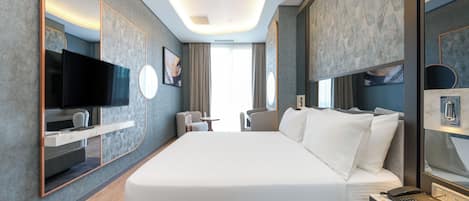 Quarto de hotel moderno com uma cama grande, televisão de tela plana, escrivaninha com telefone e área de estar com poltrona e mesa.