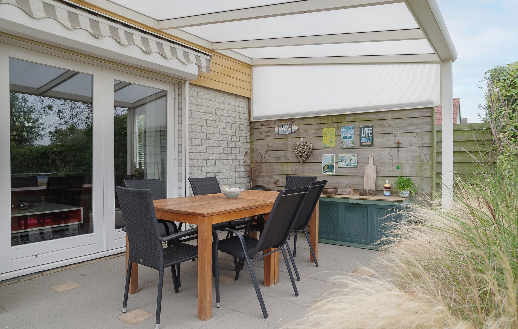Un espacio exterior para comer con una mesa y sillas de madera, una pared de ladrillo con varias decoraciones y un ventanal que da a un patio.
