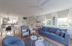 Ein modernes Wohnzimmer mit einem blauen Sofa, einem Essbereich mit Korbstühlen, einer Treppe und einer Küche im Hintergrund.