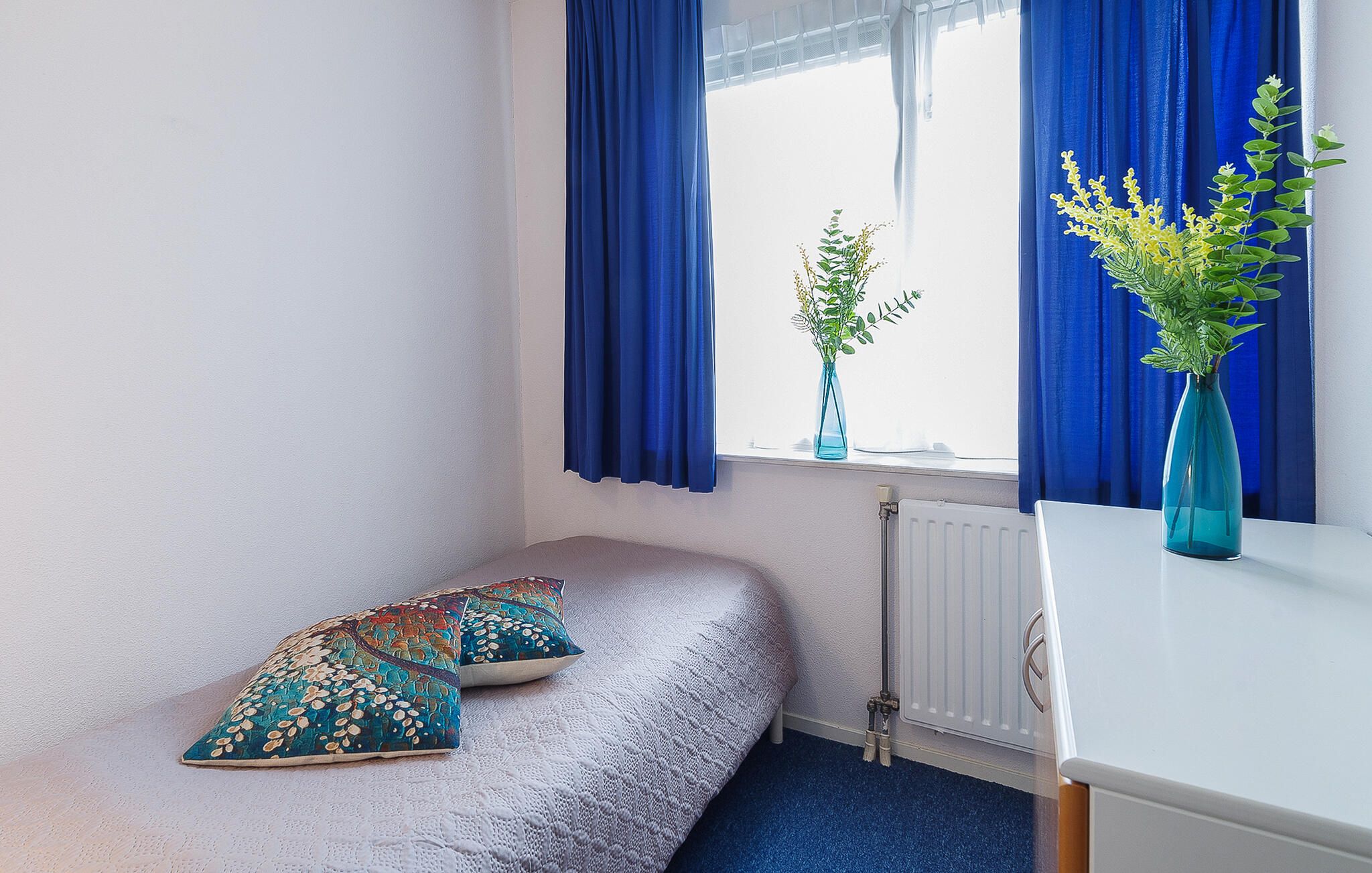 Une petite pièce avec un lit simple, deux oreillers, un radiateur et deux vases avec des plantes.