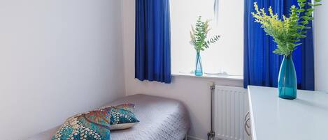 Une petite pièce avec un lit simple, deux oreillers, un radiateur et deux vases avec des plantes.