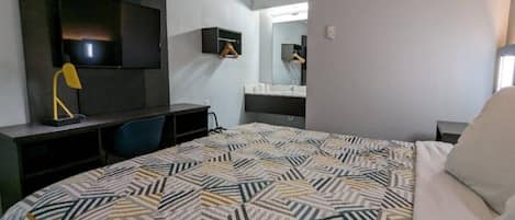 Habitación de hotel con cama, escritorio, silla, televisor, espejo y baño.