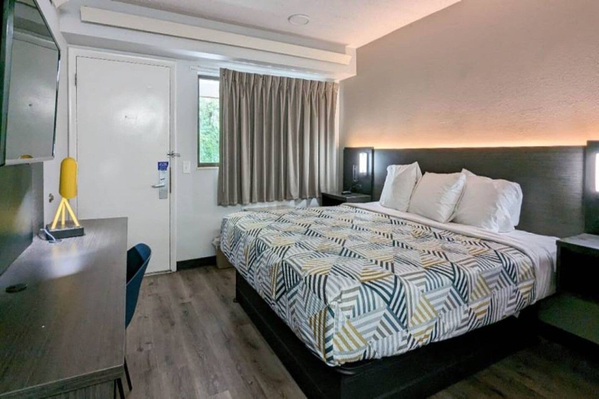 A hotel room with a bed, a desk, a chair, a window with curtains, and a wall-mounted light fixture.