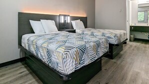 Ein Hotelzimmer mit zwei Betten, jeweils mit einem gemusterten Überwurf und weißen Kissen. Ein Nachttisch mit Lampe und Telefon ist vorhanden. Der Raum verfügt über einen Holzboden und ein Fenster mit Blick auf Grünflächen.