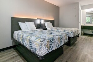 Habitación de hotel con dos camas, cada una con una colcha estampada y almohadas blancas. Se observa una mesita de noche con lámpara y teléfono. El piso es de madera y hay una ventana con vistas a zonas verdes.