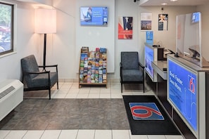 Zona de recepción con mostrador, sillas, un atril para revistas y una pantalla digital mostrando un mensaje promocional.