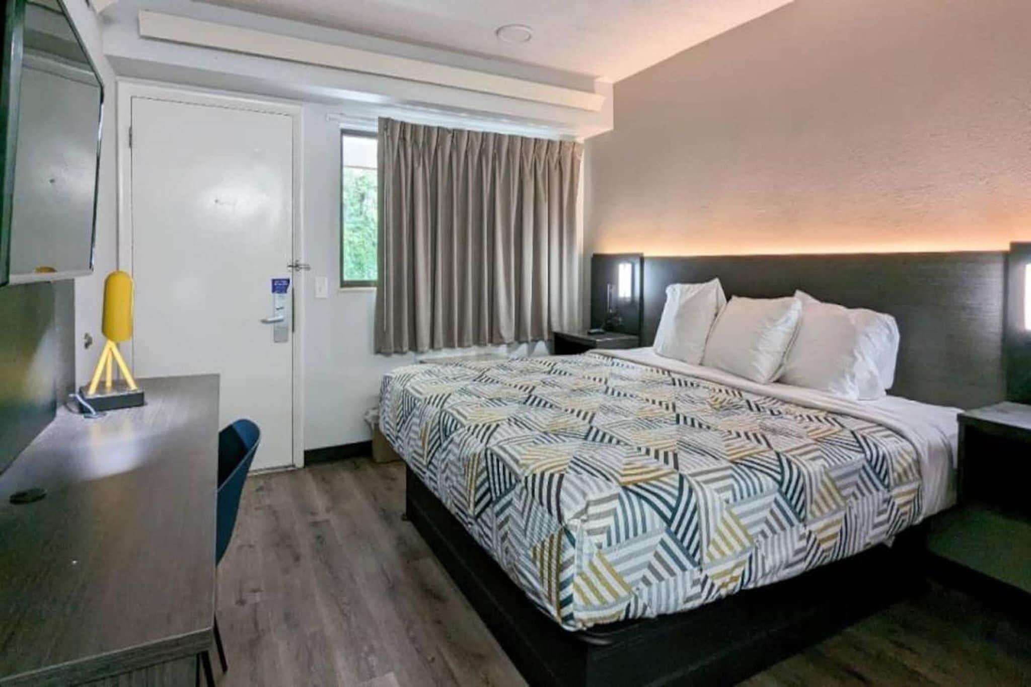 Een hotelkamer met een bed, een bureau, een stoel, een raam met gordijnen en een wandlamp.