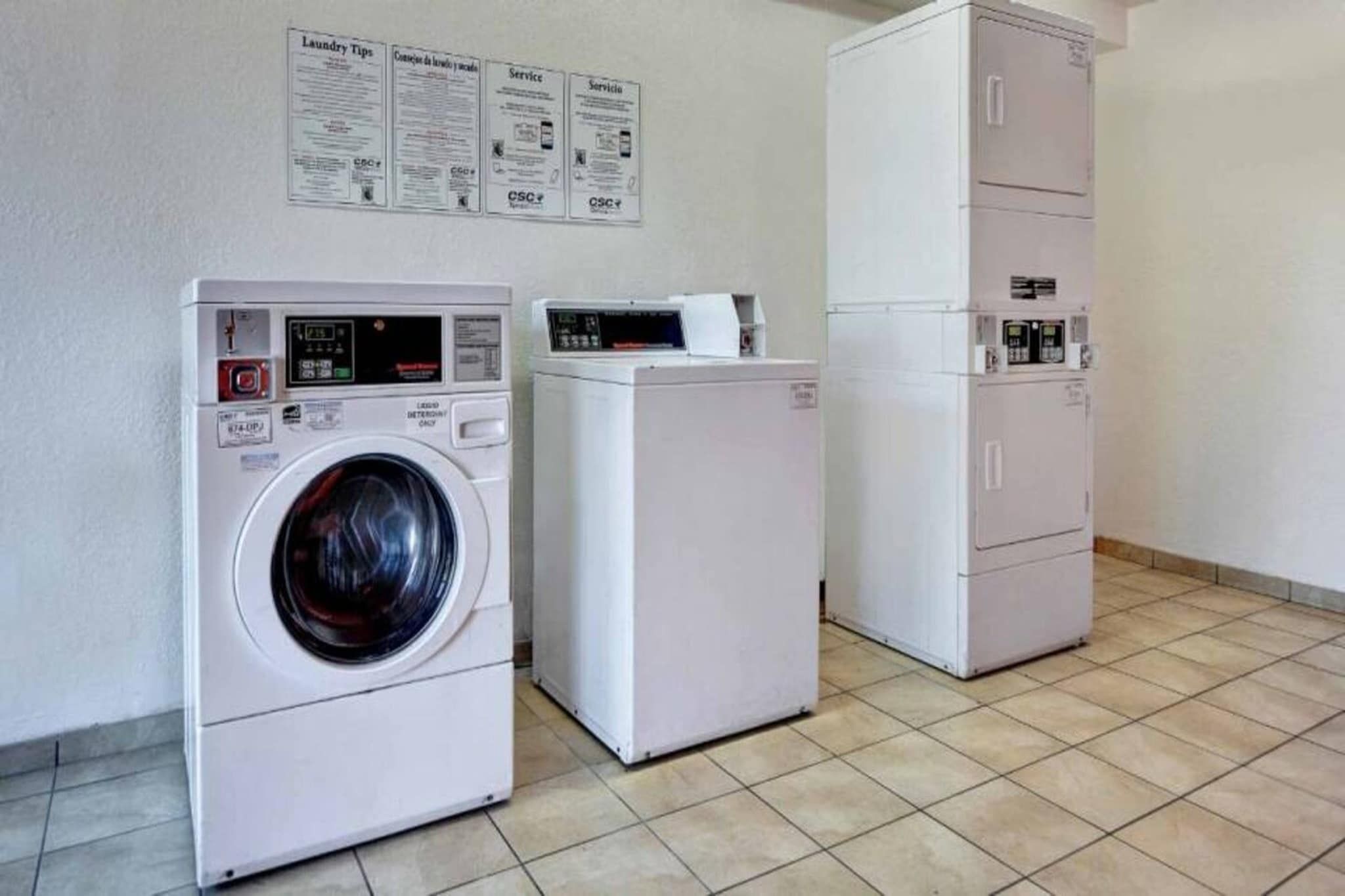 A laundry facility with a front-loading washing machine and a dryer, a cabinet with a control panel, and informational posters on the wall.