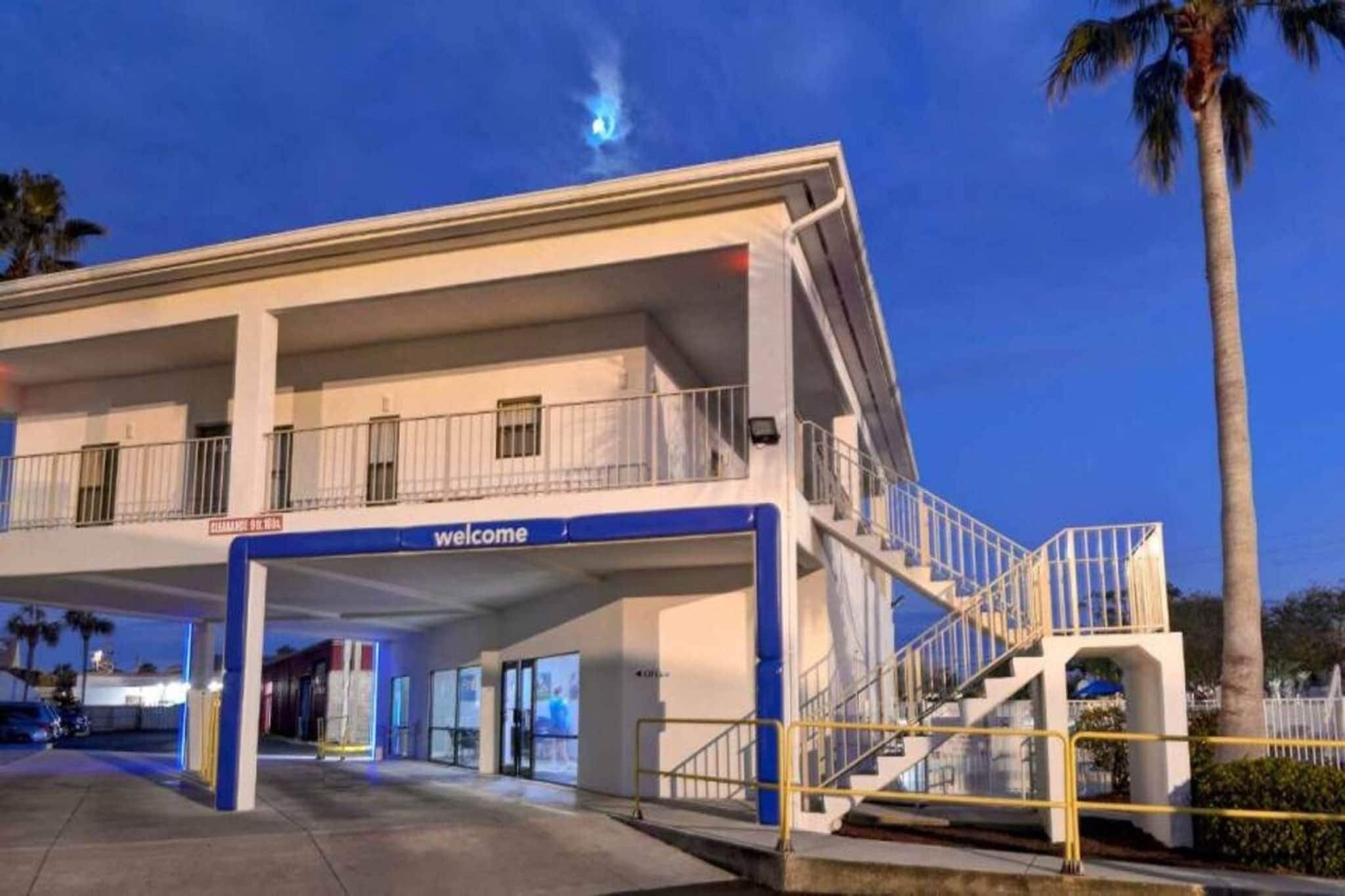 L’entrée d’un hôtel, avec une pancarte indiquant « Bienvenue », un espace de stationnement couvert et un escalier menant à l’étage supérieur.