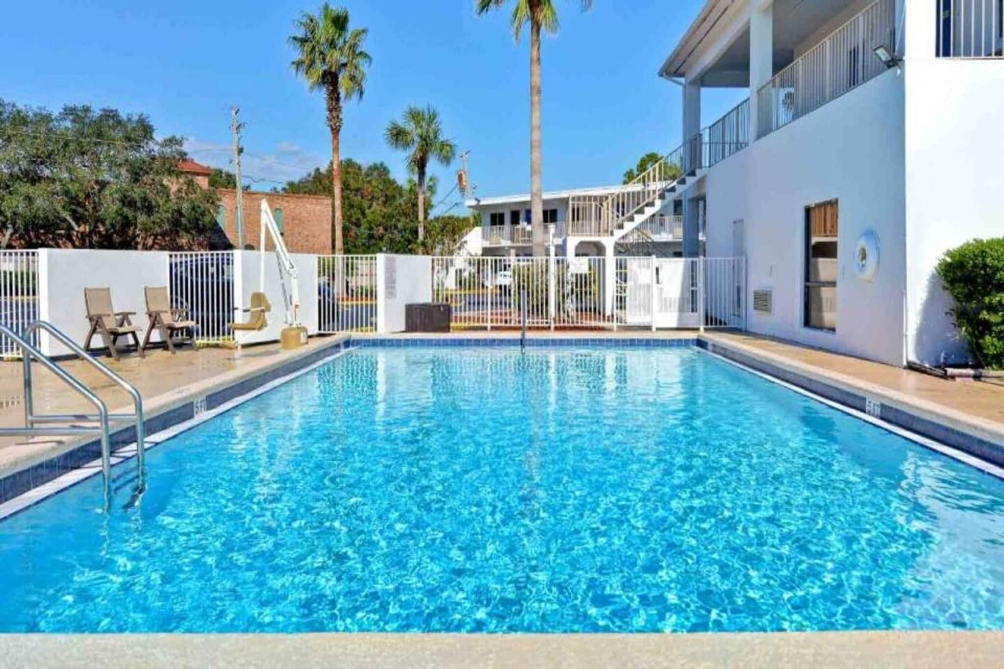 A rectangular swimming pool with a clear blue water surface, surrounded by a white fence, and adjacent to a building with a balcony.